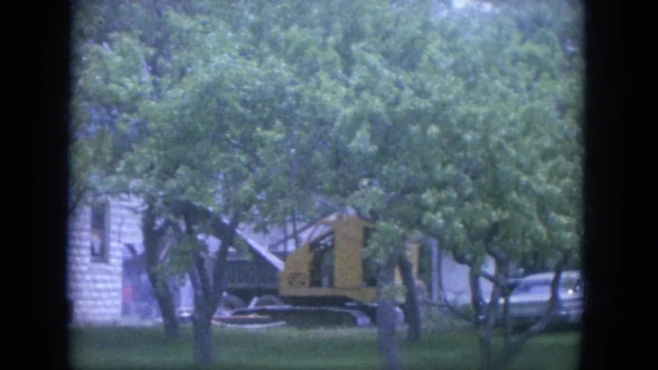 Louisville 1965 home-movie frame showing a tree-lined yard, yellow construction equipment, a parked car, and residential buildings.