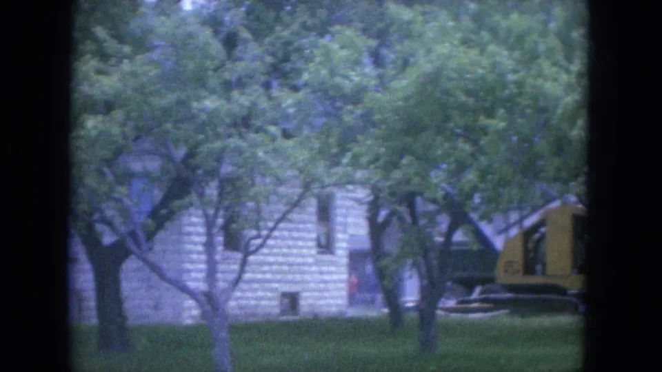 1965 Louisville residential view through a window frame, showing trees, grass, a house facade, and yellow equipment at right.