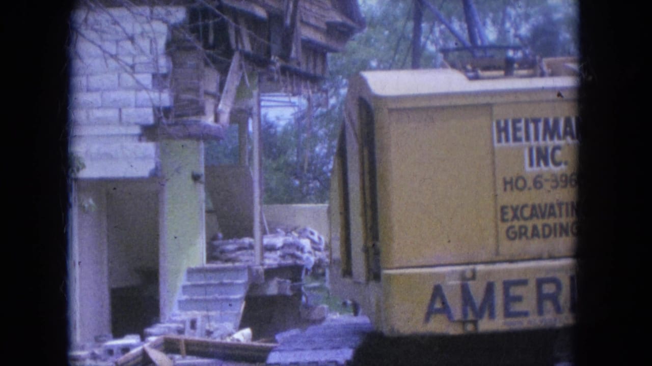 Yellow excavator beside a partly demolished house with porch columns and debris.