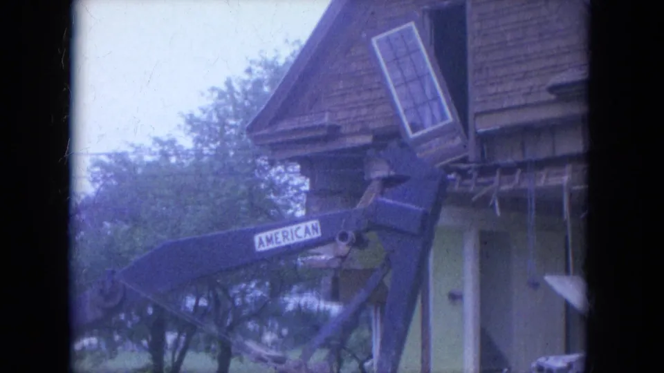 1965 Louisville 8mm frame showing an AMERICAN-marked excavator boom raised against a wood-frame house with a tilted window and scattered debris.