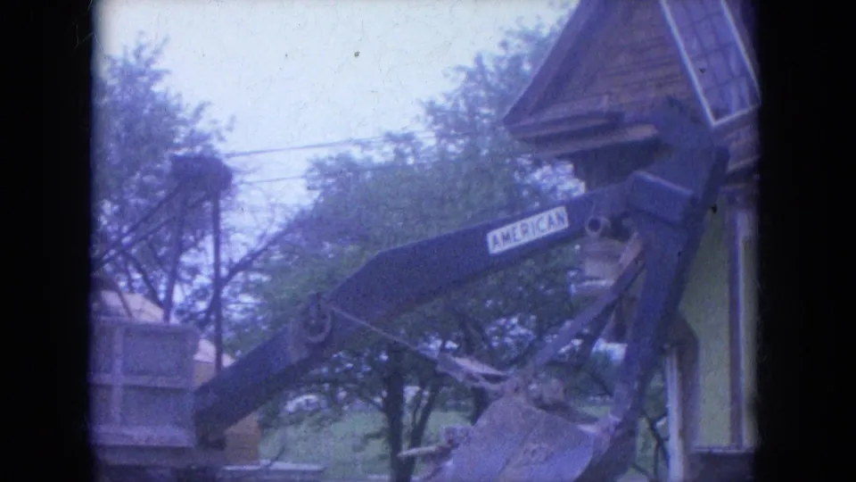 1965 Louisville home-movie frame showing the AMERICAN-marked excavator arm and bucket near a house corner, with trees and a pale sky behind.