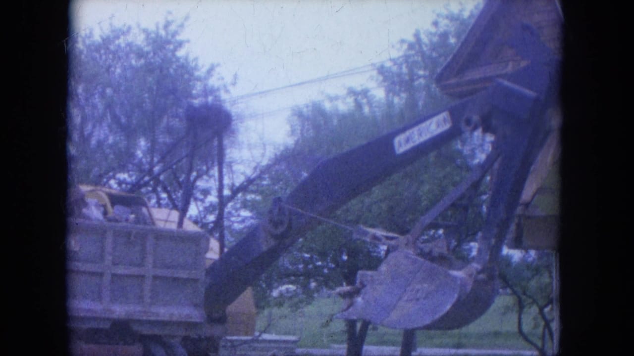 1965 Louisville 8mm frame showing an excavator arm and bucket loading debris near a dump truck, with trees and overhead lines in view.