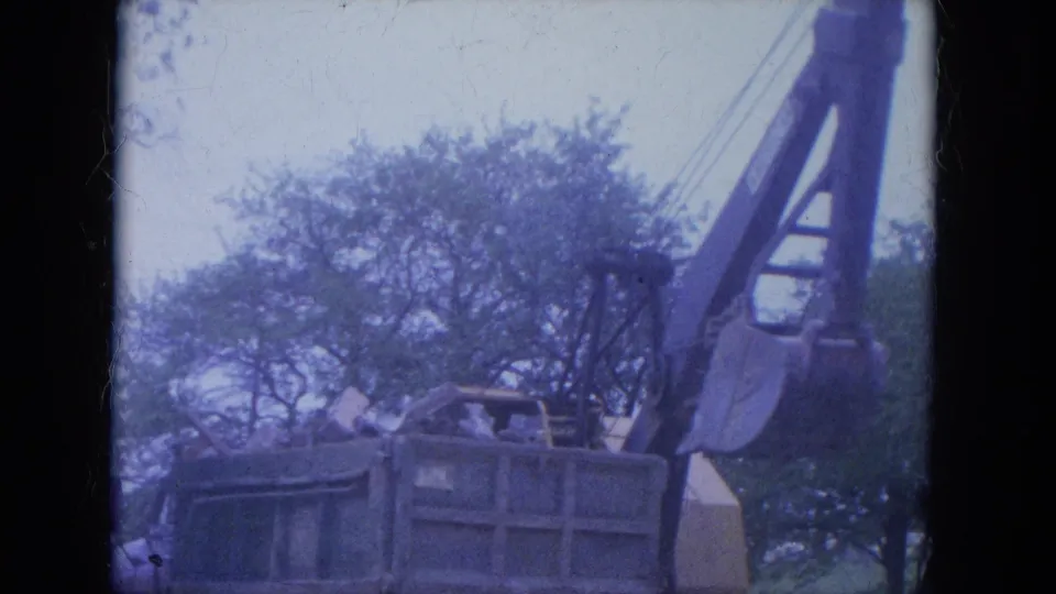 A dump truck sits below the raised excavator bucket as the home-movie camera frames the machinery against trees and sky.