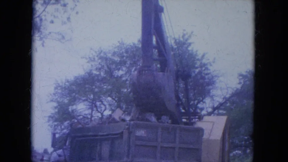 The excavator bucket hangs above the dump truck near the end of the 1965 Louisville construction clip, with trees and pale sky behind.