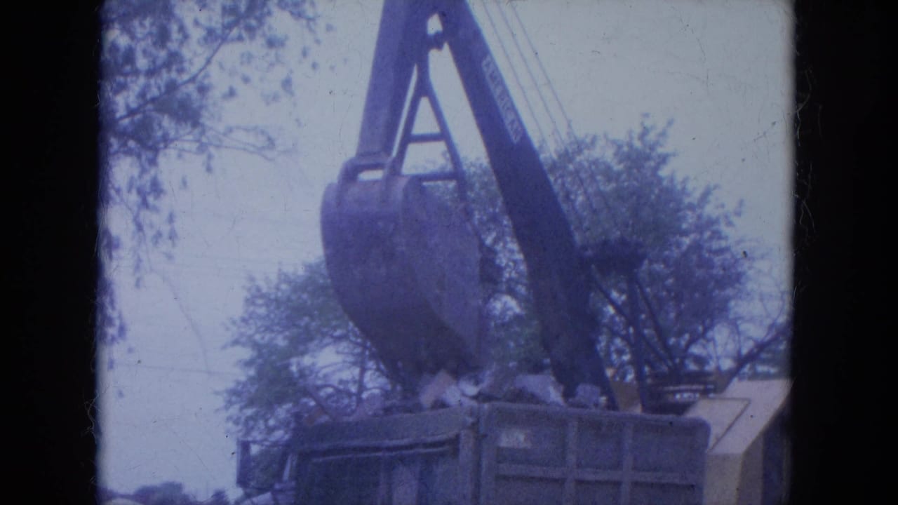 Crane bucket over a debris container beside trees and a damaged building edge.