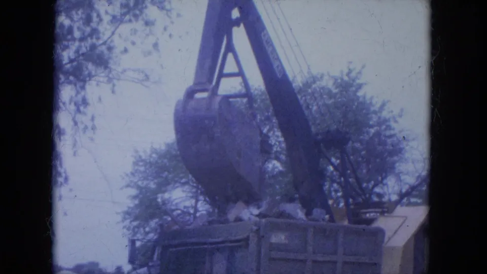 1965 Louisville 8mm frame showing a crane bucket positioned above a debris container near trees and a damaged house exterior.