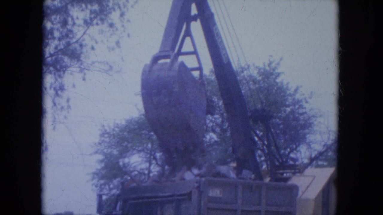 Crane bucket and boom above a container with trees in the background.