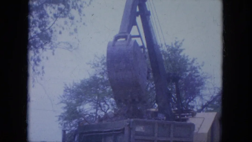 Home-movie frame from the Louisville demolition scene showing the crane bucket lowered near a debris container beneath trees.