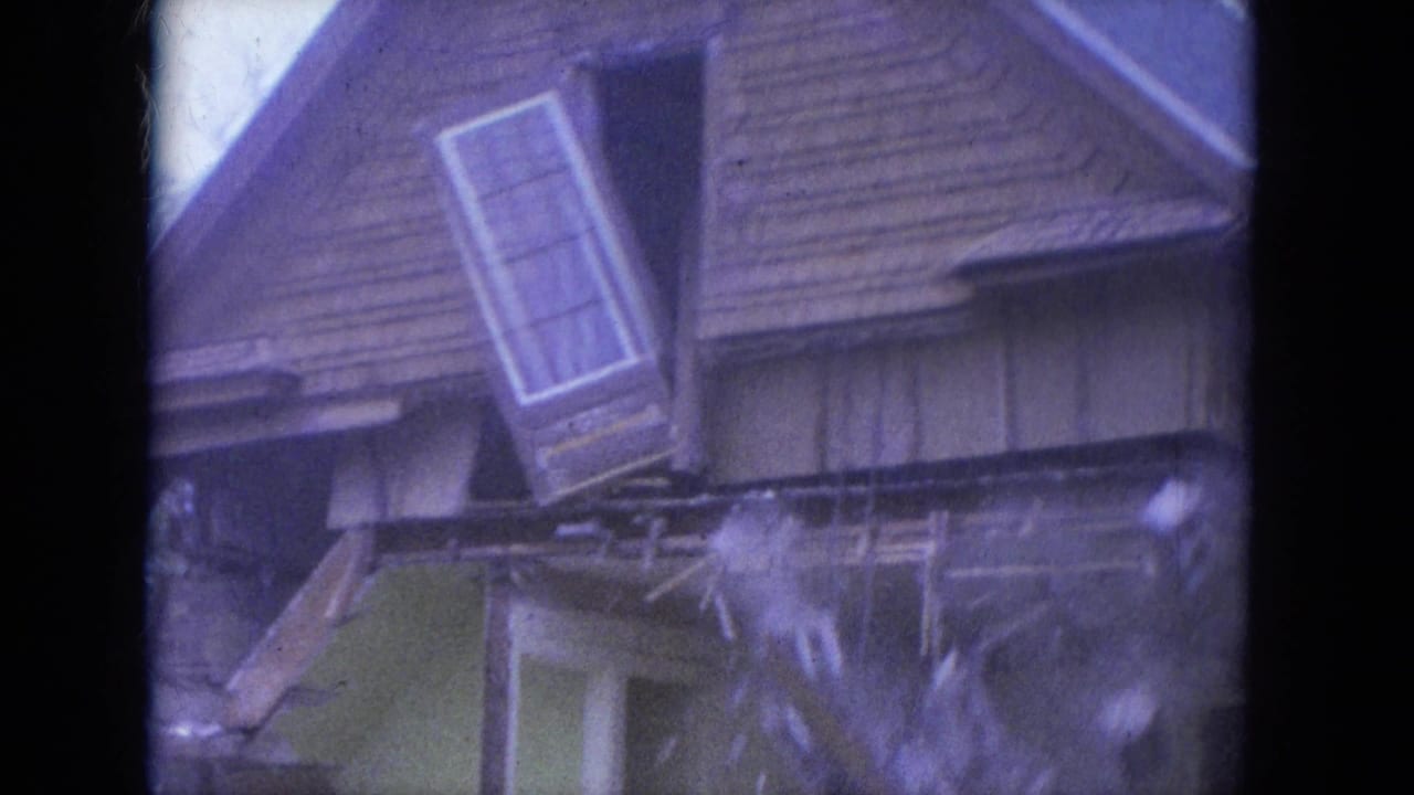 Damaged upper story of a wood-sided house with a loose window assembly hanging near the roofline.
