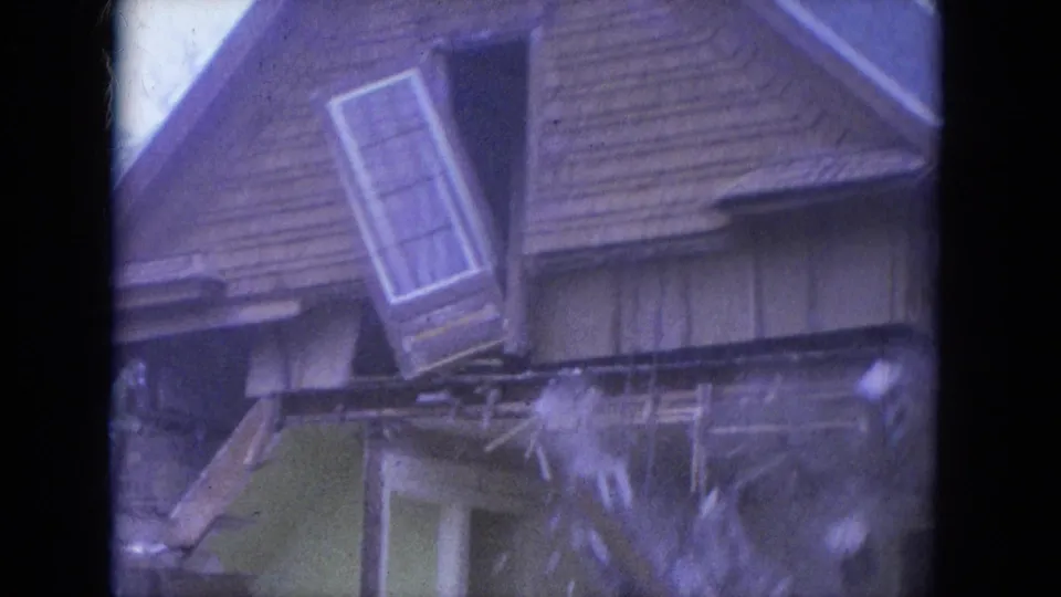 1965 Louisville home-movie frame showing a steep shingled roof, broken upper framing, and a dislodged window or door assembly hanging outward.