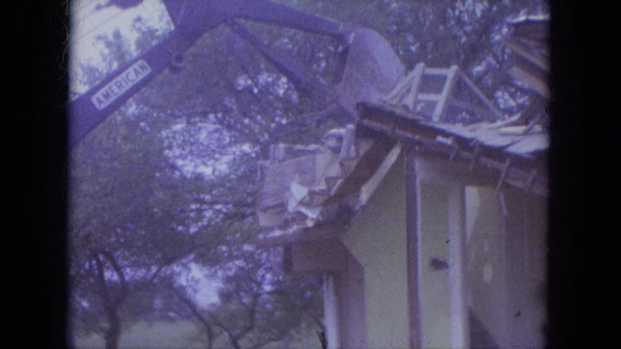 Excavator arm marked AMERICAN reaches over the roof of a small house among trees.