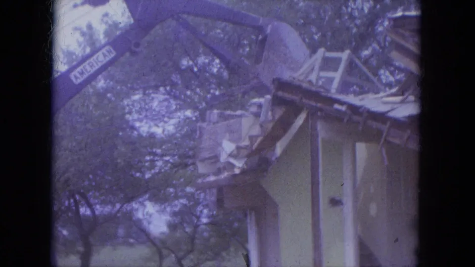 1965 Louisville demolition frame with an excavator arm marked AMERICAN positioned above a small house roof under trees.