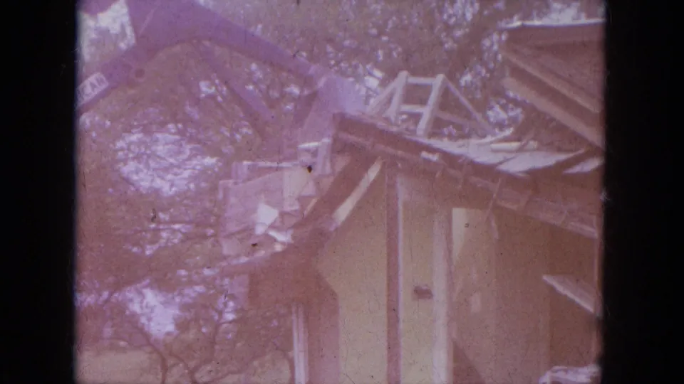 Faded 1965 Louisville 8mm frame showing an excavator working over a small house with torn roof boards and trees behind it.