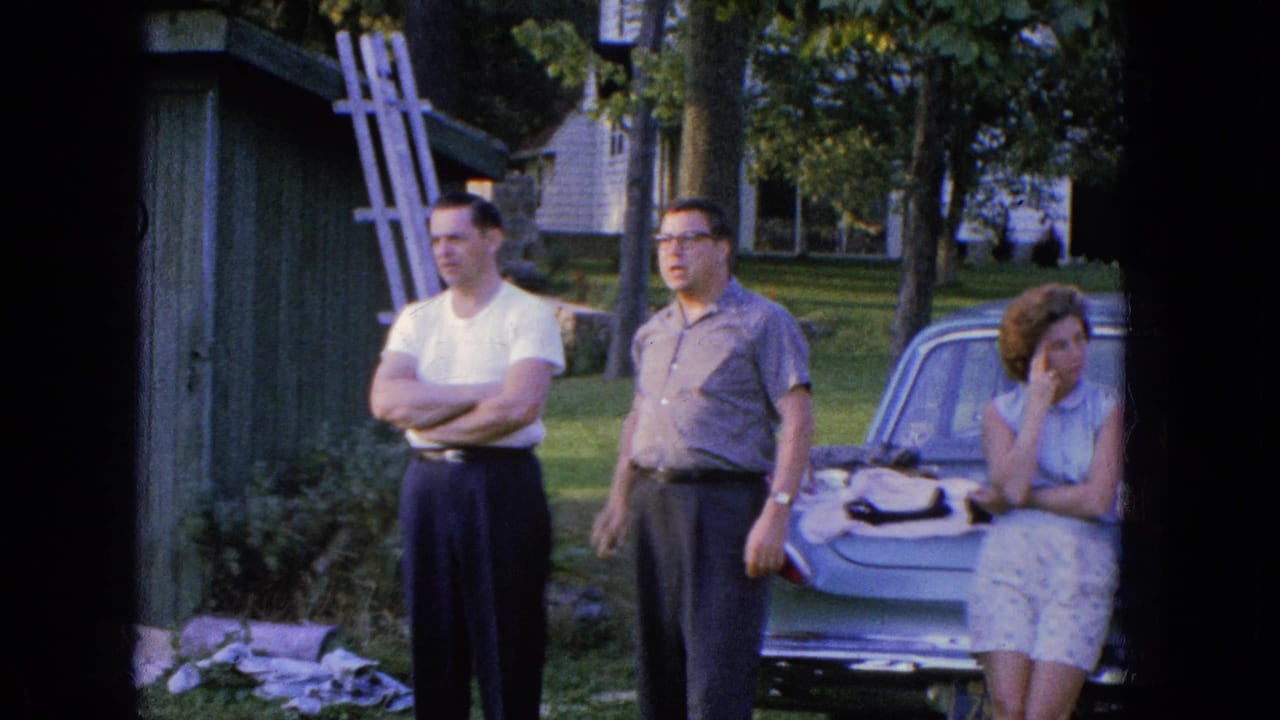 In Manchester, New Hampshire in 1965, two men stand by a parked car as a woman sits on the trunk near a green shed and trees.