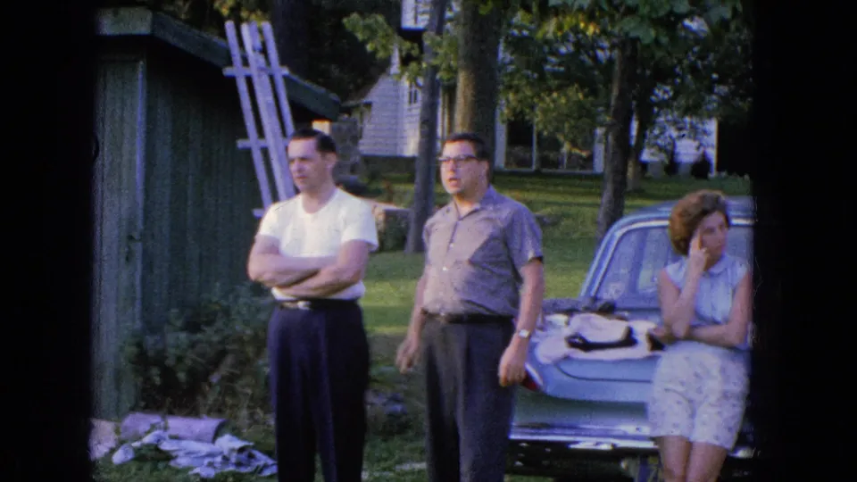 In Manchester, New Hampshire in 1965, two men stand by a parked car as a woman sits on the trunk near a green shed and trees.