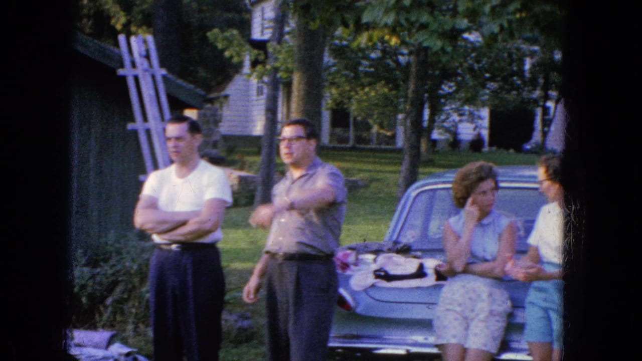 In the 1965 Manchester home-movie scene, adults converse around a parked car, with one man gesturing and two women near the trunk.