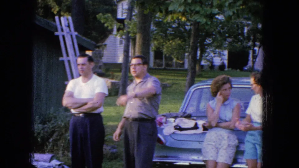 In the 1965 Manchester home-movie scene, adults converse around a parked car, with one man gesturing and two women near the trunk.