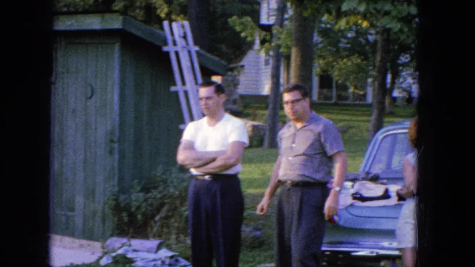 Two men stand in a grassy Manchester yard in 1965, with a parked car and a partly visible seated woman near the frame edge.