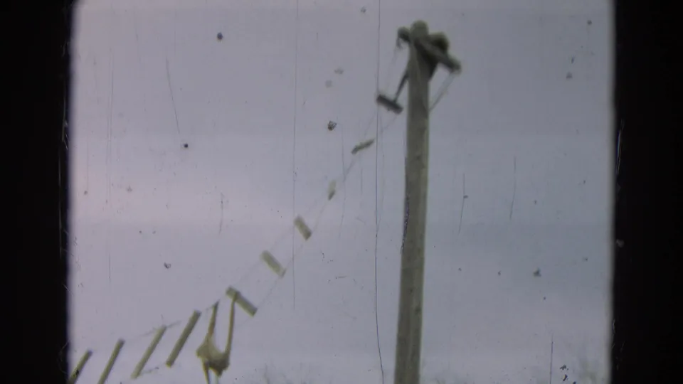 1965 Omaha home-movie frame showing a wooden post, rope supports, and suspended bridge slats against a pale sky.