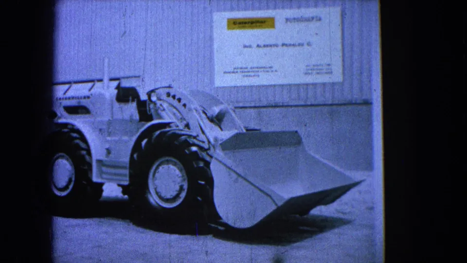 1965 8mm frame showing a white Caterpillar-style front-end loader in a warehouse or showroom setting with signage on the wall.