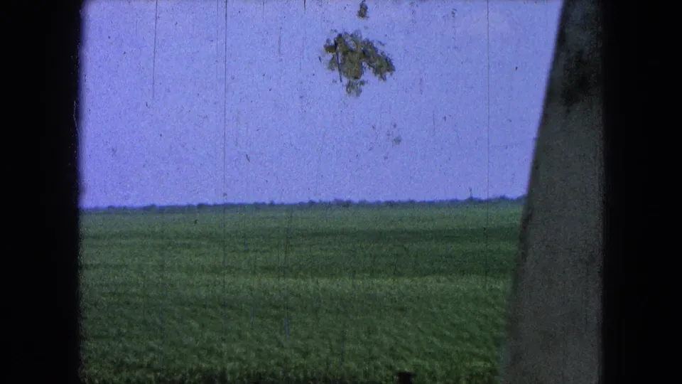 Pueblo, Mexico home-movie frame showing a green field, pale sky, dark side obstruction, and visible 8mm surface marks.