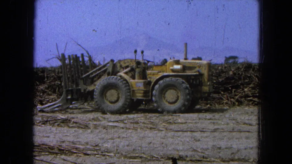 Yellow agricultural vehicle parked in a harvested Pueblo, Mexico field, with stalk piles, distant hills, blue sky, and dark 8mm edges.