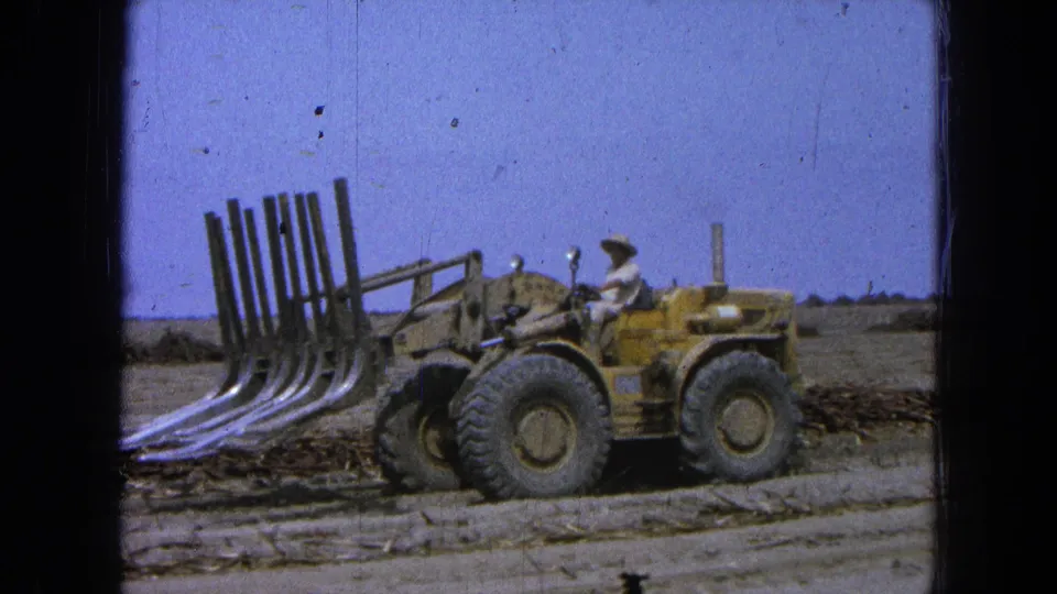 1965 Pueblo, Mexico 8mm frame showing a driver in a light shirt and hat operating a yellow loader with raised tines.