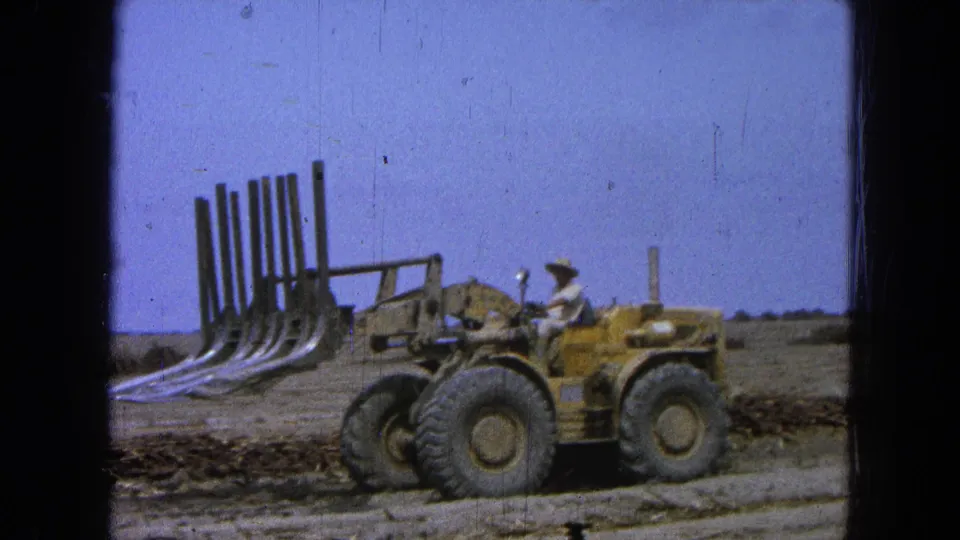 1965 Pueblo, Mexico frame showing a man seated on a yellow loader with a large grapple attachment beside brush and open land.