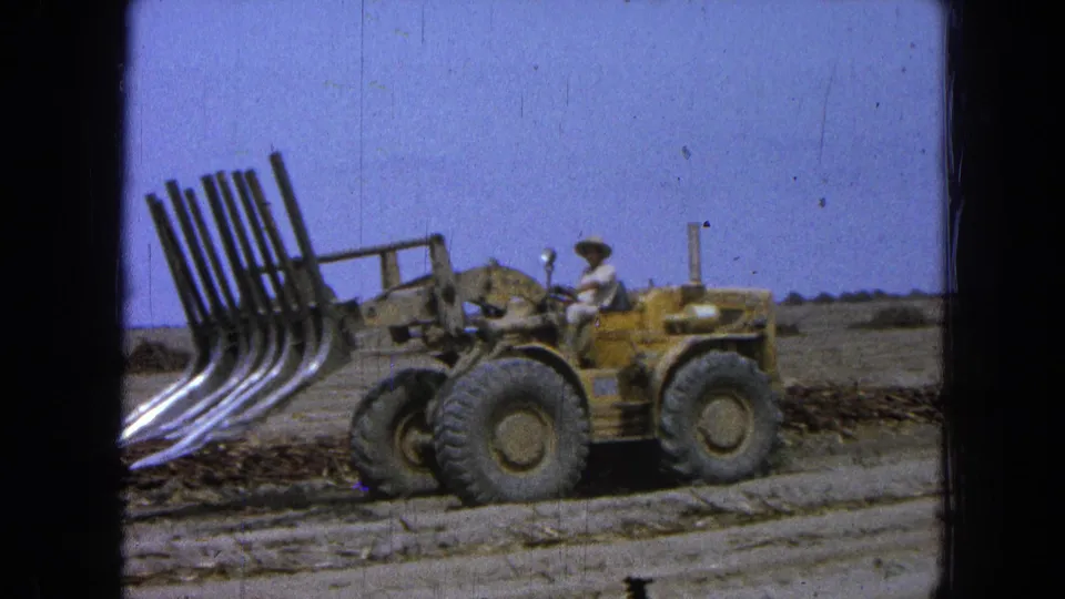 1965 Pueblo, Mexico frame with the loader in profile, its curved grapple tines raised near a dirt roadway and cut brush.