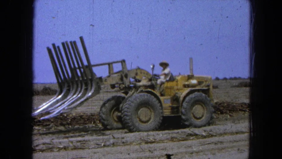 1965 Pueblo, Mexico frame showing a man driving a yellow loader with a front grapple past scattered brush on a dusty rural road.