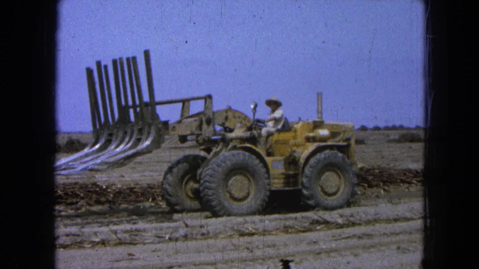 1965 Pueblo, Mexico frame showing the loader and operator beside cut brush, with open ground and a clear sky in the background.