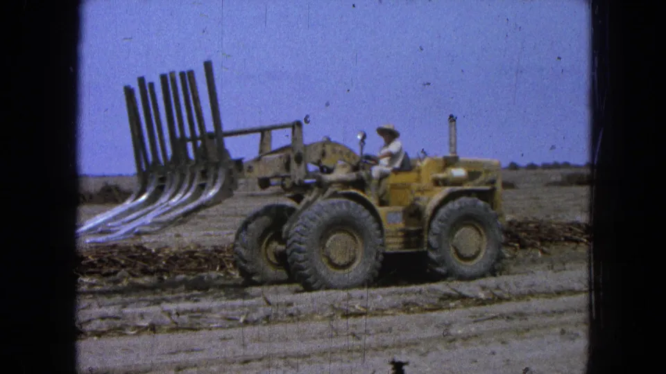 In supplied 1965 Pueblo, Mexico footage, a man drives a yellow front-end loader with raised curved tines across dry open ground.