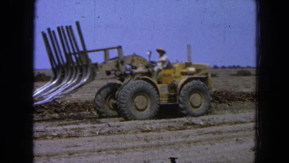 The yellow loader appears in side profile with its seated operator and raised tine attachment against a clear sky and flat field.