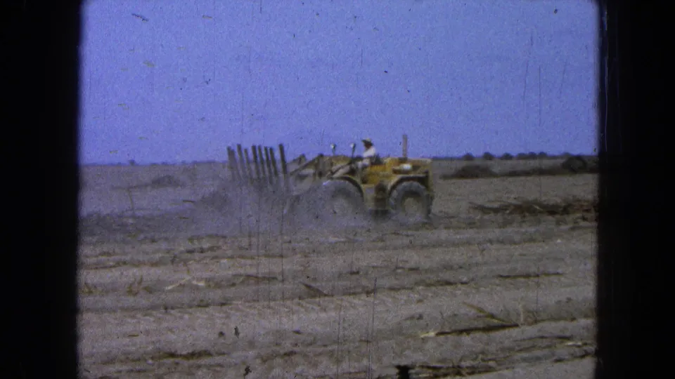 A yellow tractor continues field preparation in rural Pueblo, Mexico, with the driver visible against the dusty landscape.