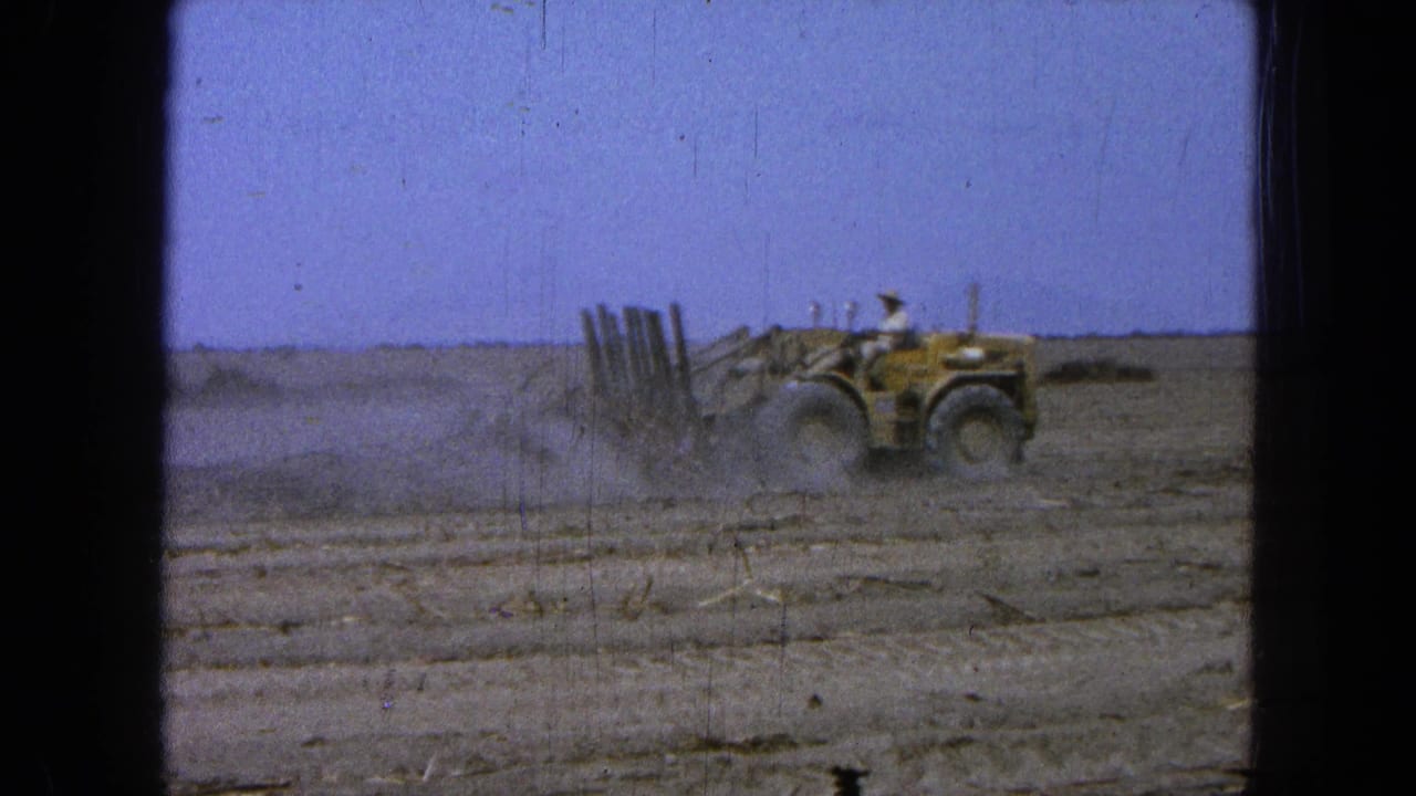 Yellow tractor moving across a dry field, raising dust under a blue sky.