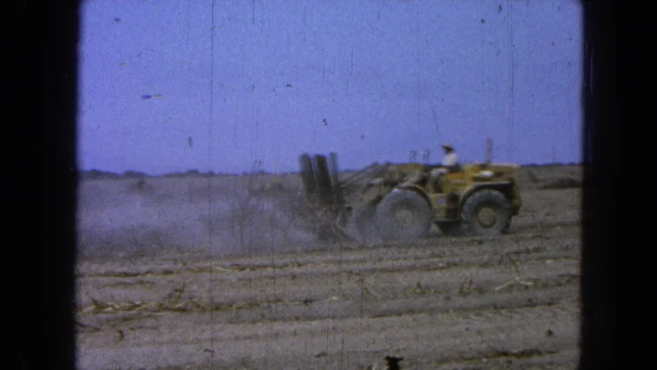 The 8mm frame shows the tractor clearing dry ground, with the operator small in view and dust spreading behind.
