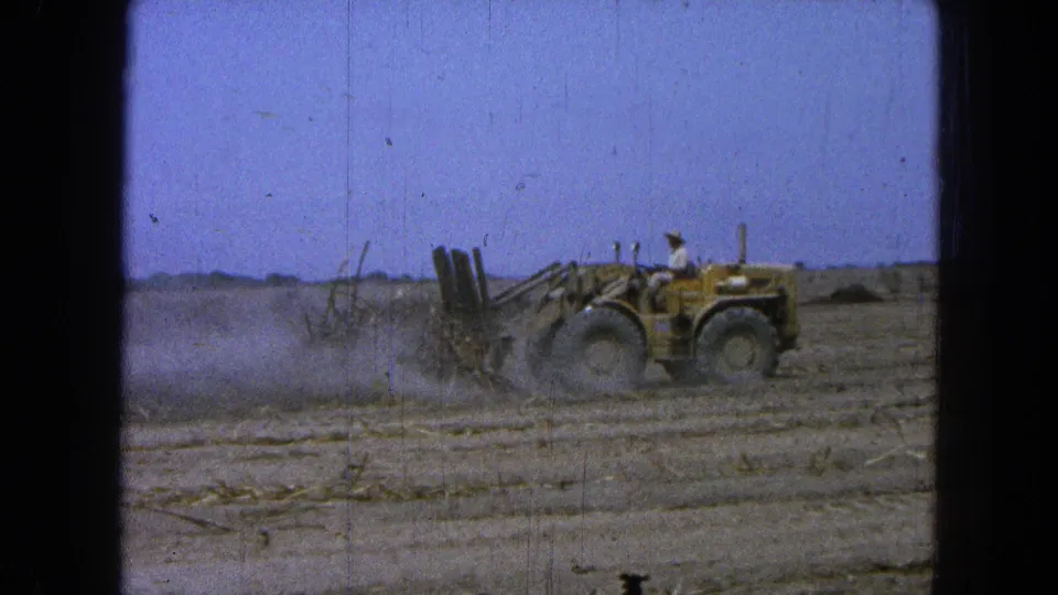 A tractor operator clears a dry rural field in 1965 Pueblo, Mexico, framed by blue sky and visible 8mm film wear.