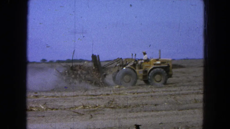 The loader advances through a dry Pueblo, Mexico field, with the operator visible and dust spreading behind the pushed debris.