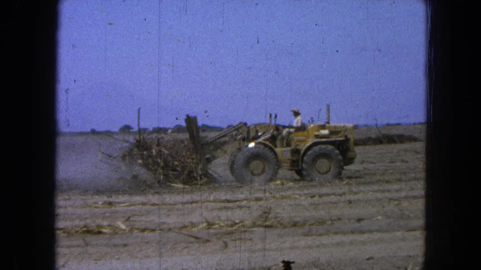 A yellow loader with a visible operator pushes brush and dirt through a dusty field in archival 8mm footage from Pueblo, Mexico.