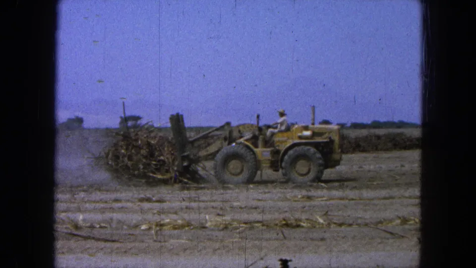 1965 Pueblo Mexico 8mm frame showing a yellow front-end loader with a driver pushing agricultural debris in a dry field.
