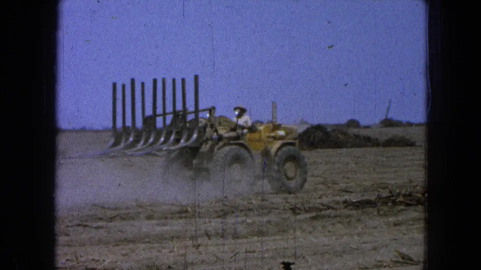 A worker in the 1965 Pueblo, Mexico footage drives a yellow loader across a dry field, passing scattered crop debris and low dust.