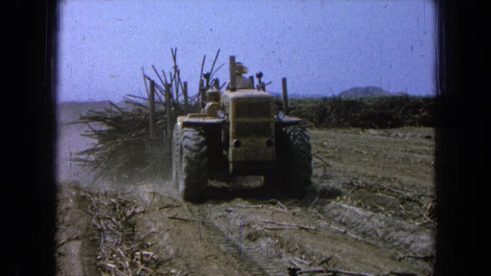 Final frame of the yellow tractor working through dusty terrain, with pulled brush and low hills visible in the background.