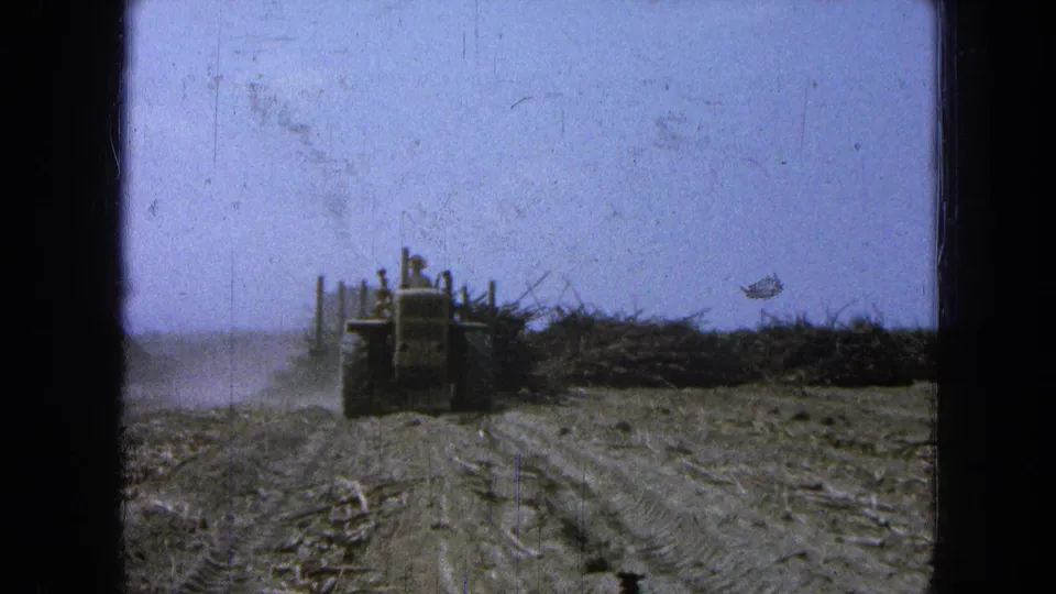 The 8mm home-movie view shows the tractor farther away on a dusty field road in Pueblo, Mexico, with crop debris and a flat horizon.