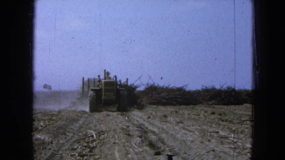 A distant tractor continues away from the camera on a dusty Pueblo, Mexico farm road, framed by visible 8mm film scratches and borders.