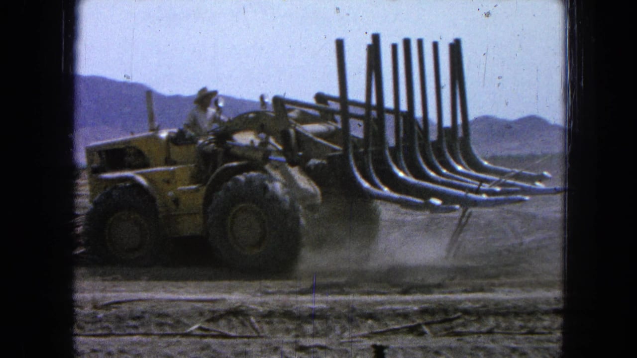 Driver in a wide-brimmed hat operates a yellow loader with raised grapple arms.