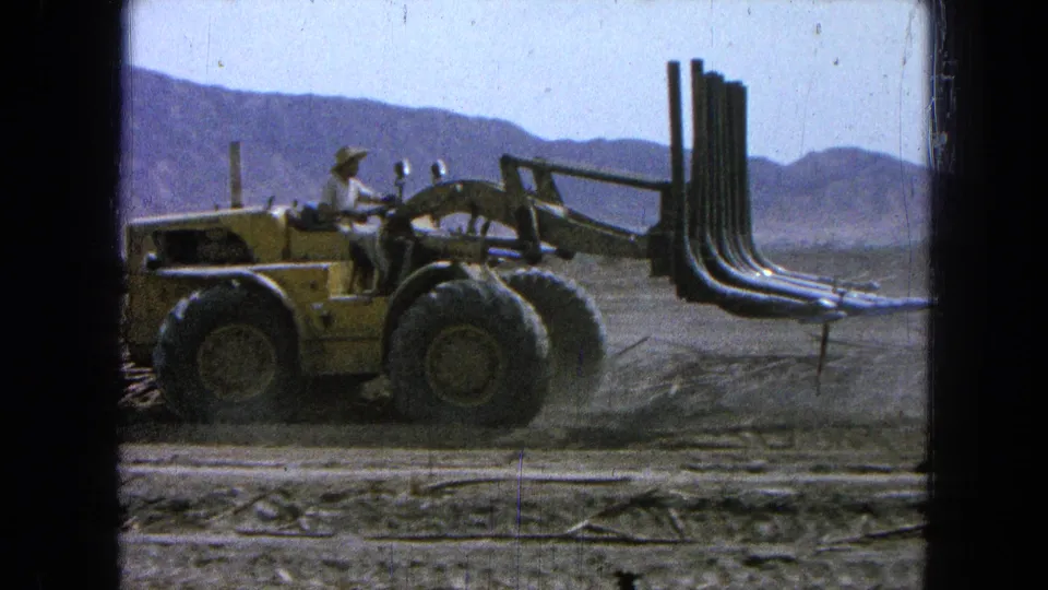 The yellow loader moves side-on across the field, its grapple attachment extended as mountains sit in the background.