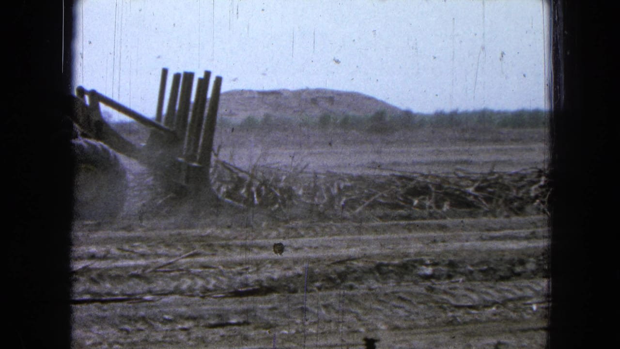 Front-end loader pushes debris across a dusty field behind dark window framing.