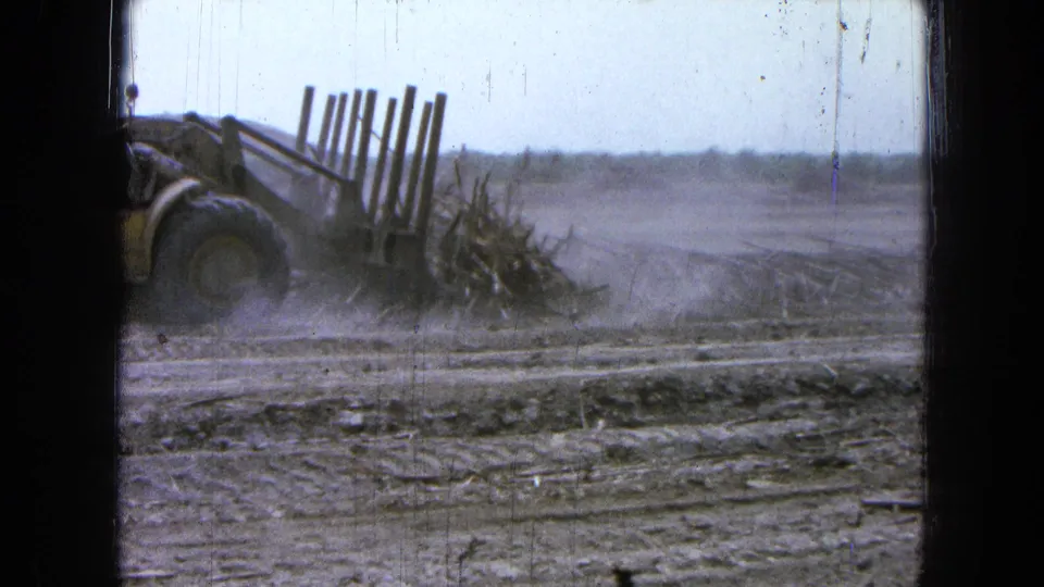 A yellow loader continues clearing debris in the dusty 1965 Mexico field, with the machine partly cut off by the left frame edge.