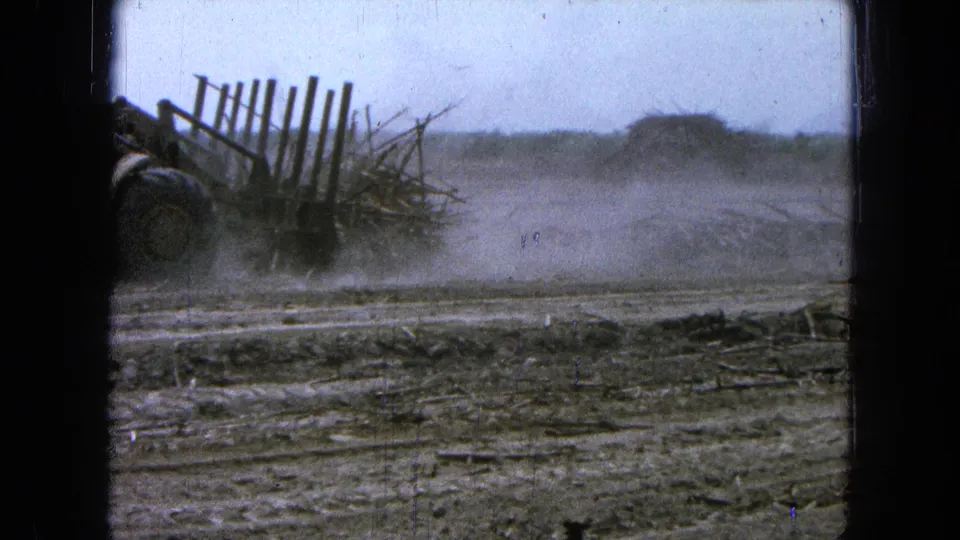 1965 Pueblo, Mexico home-movie frame showing a tractor at the left edge pulling a multi-blade plow through dust in a dry field.