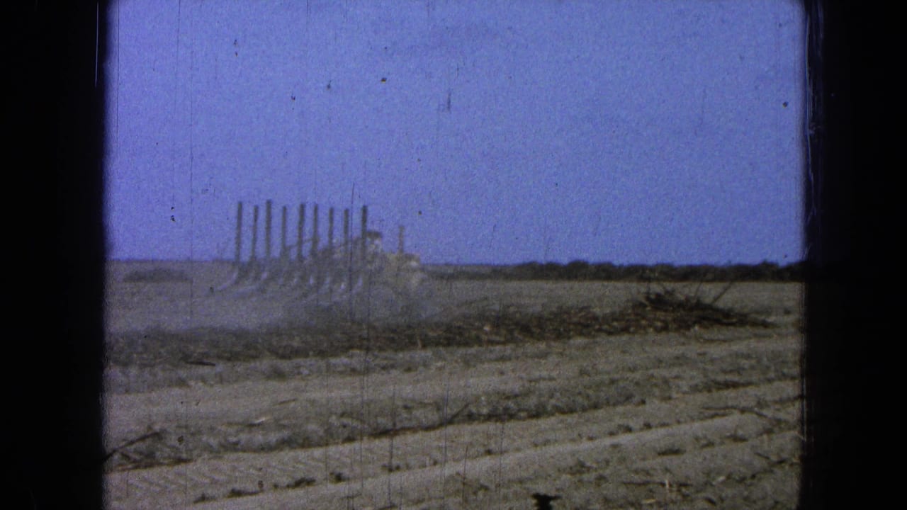 1965 Pueblo, Mexico frame with the plow farther across the field, dust drifting around its blades beneath a clear blue sky.